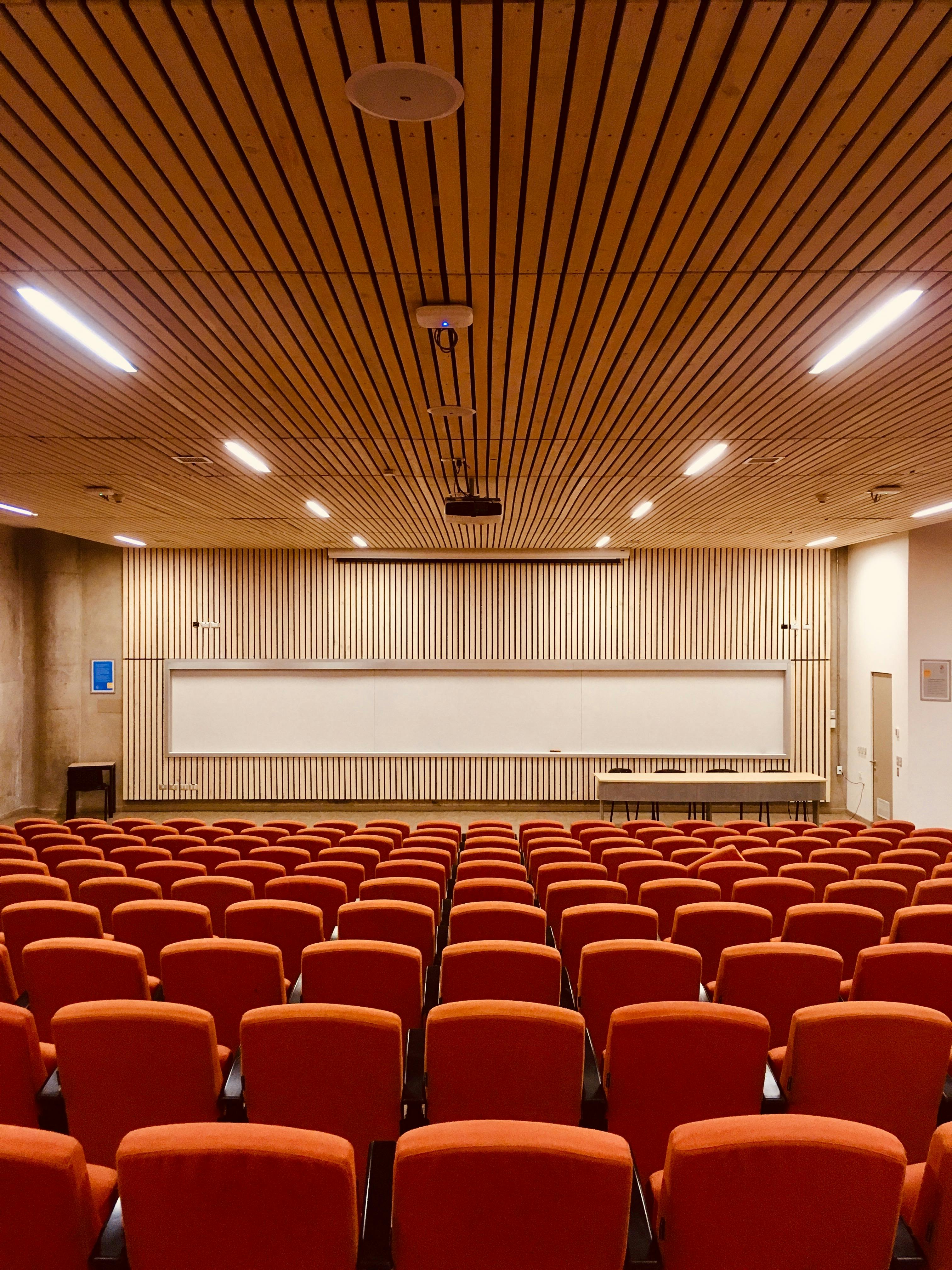 Bright, cozy and colorful image of an empty classroom. The bold, brick-colored chairs form an interesting texture in the bottom one third of the image, contrasting with the light tones of the wooden wall at the back of the room. Competing with the intentional symmetry of the image, one of the chairs at the bottom is slighty angled.