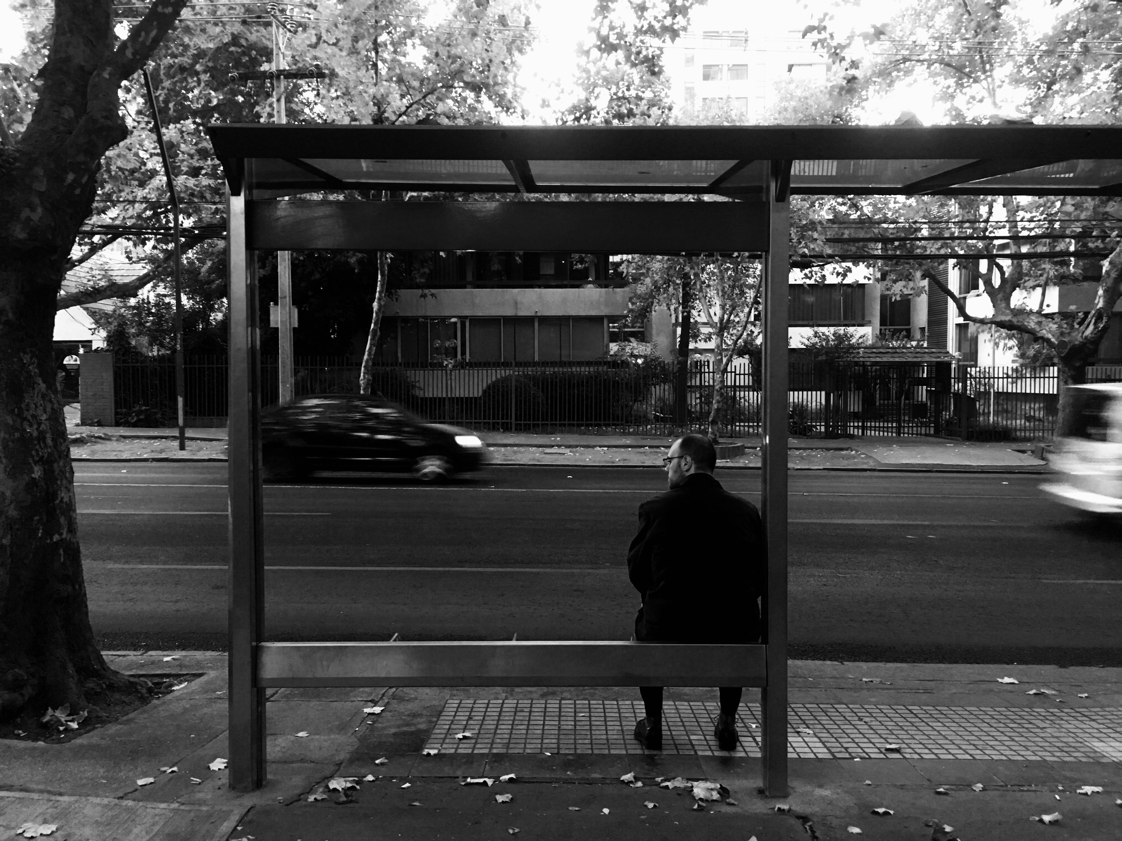 Black and white image of a man waiting alone in a bus stop while cars pass by in the back. The cars are motion-blurred, and the man seems to be looking at the photographer suspicously.