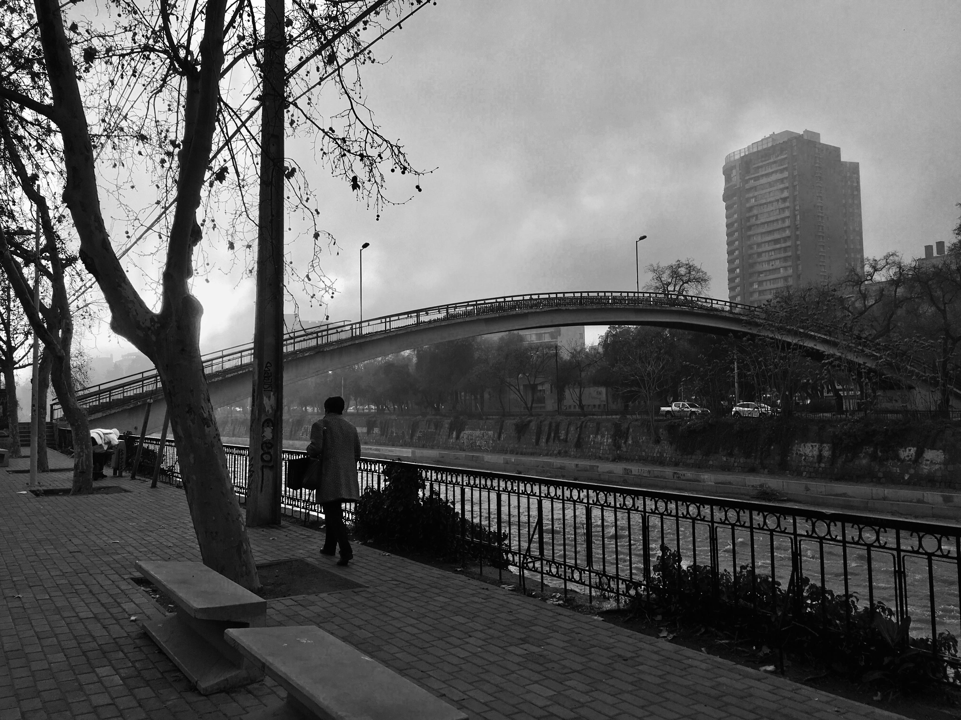 Low contrast, black and white image of a woman walking by the Mapocho river. The day is cloudy and the sidewalk is empty. The lighting of the image gives a nostalgic and melancholic feel.