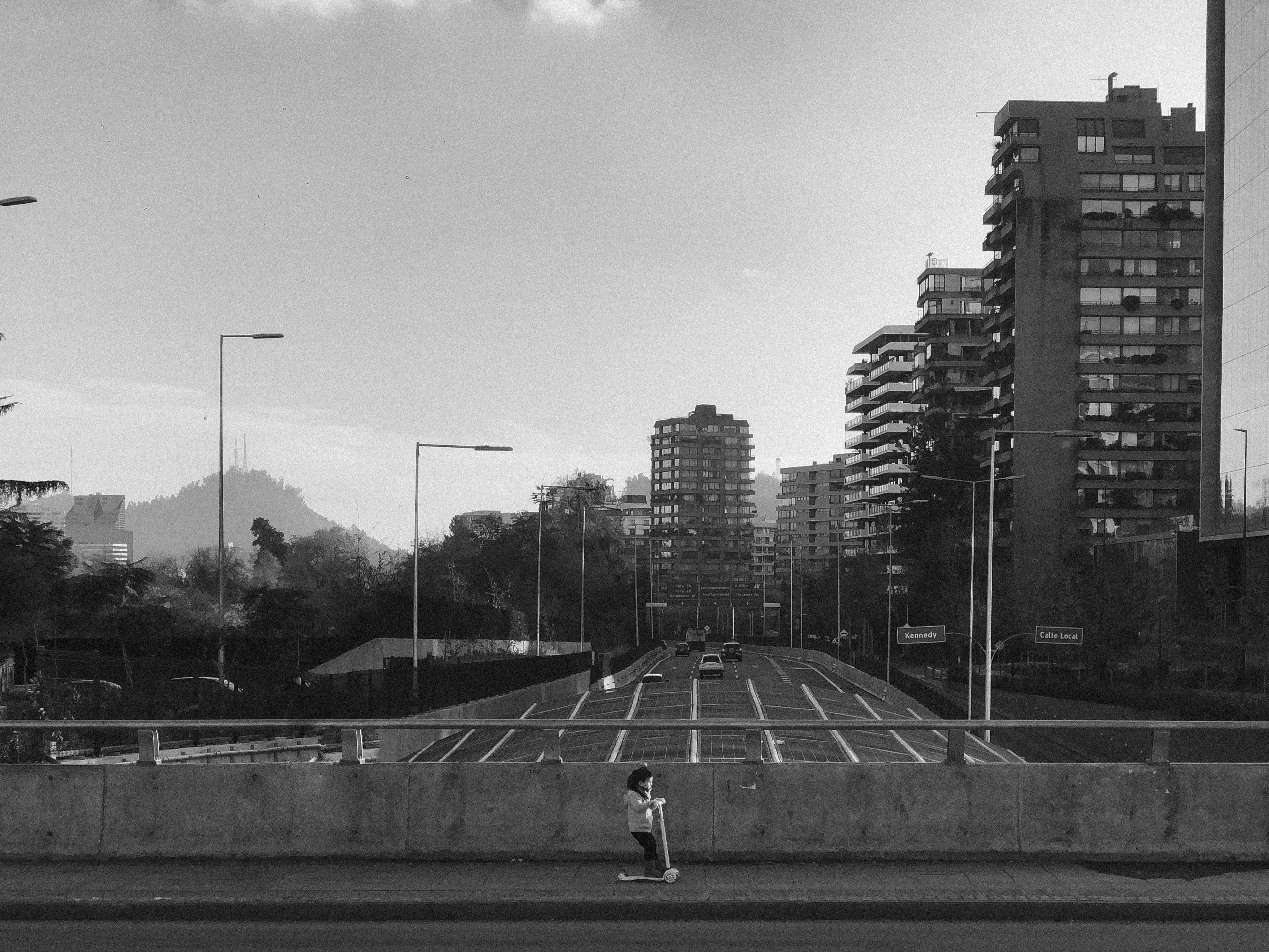 Black and white picture of a small child riding a miniature scooter, alone, through a massive highway. Noisy, low-contrast image.