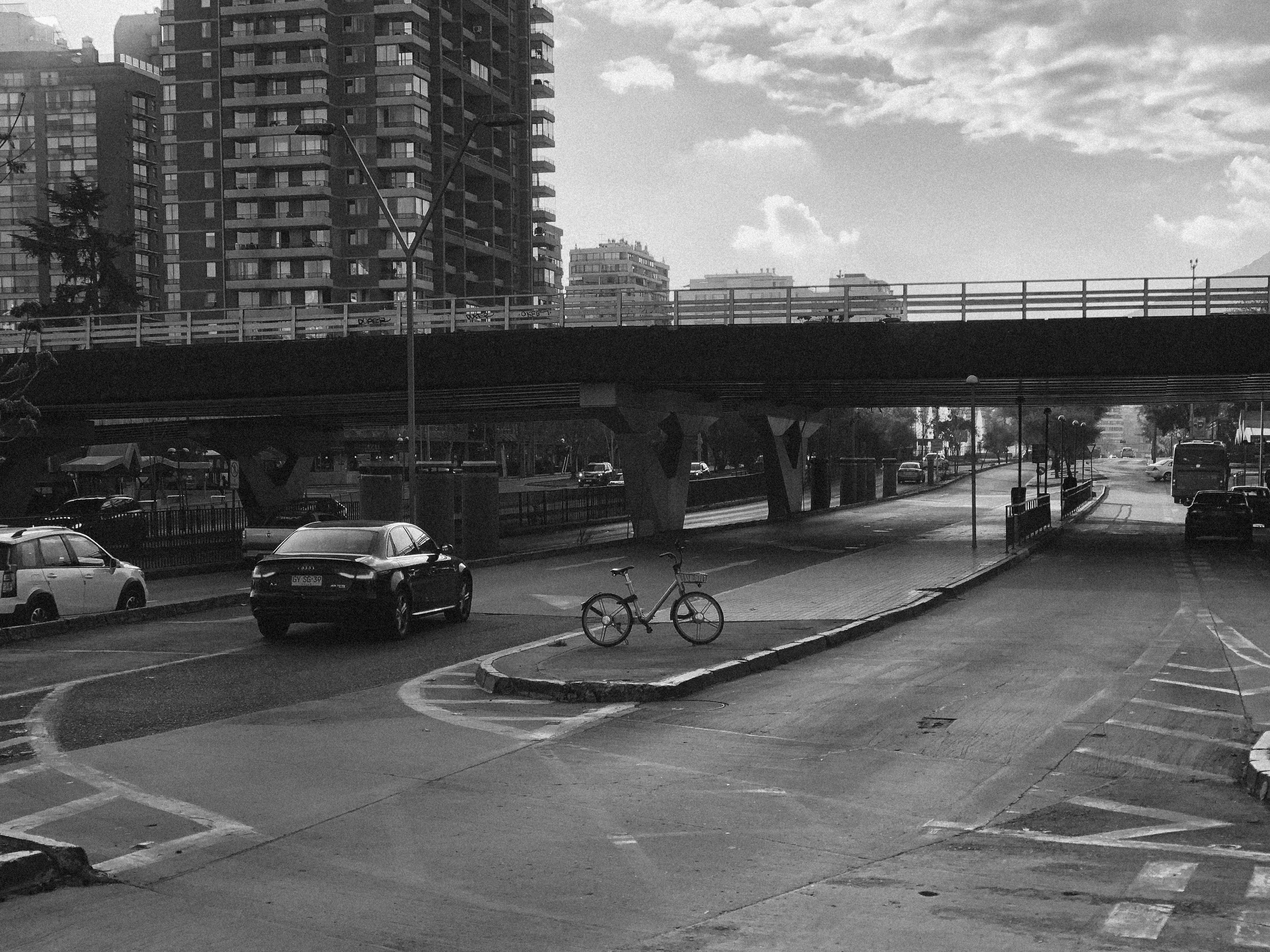 Black and white picture of a shared bicycle left alone in a small concrete island between car lanes. Noisy, low-contrast image.